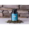 Een flesje MindScopic - CLEAR FOCUS™ Cafeïnevrije Capsules (50 stuks) rust op een wit oppervlak met losse kruidenbladeren in de buurt en een stenen muur op de achtergrond. miniatuur