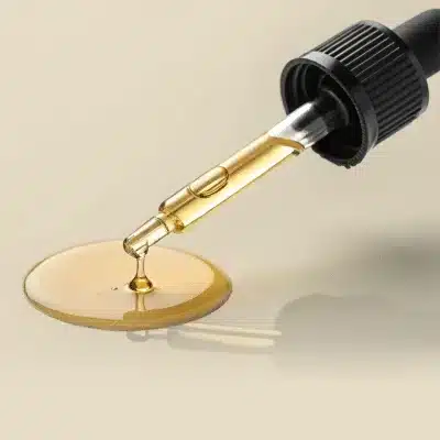 Een druppelaar die een goudkleurige vloeistof op een glad, licht oppervlak spuit, waarbij zich een klein plasje vormt onder de punt van de druppelaar.