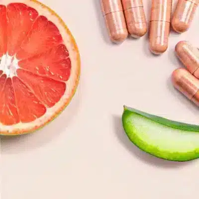 Een schijfje grapefruit, een stukje aloëvera en een aantal koperkleurige capsules liggen op een licht oppervlak.